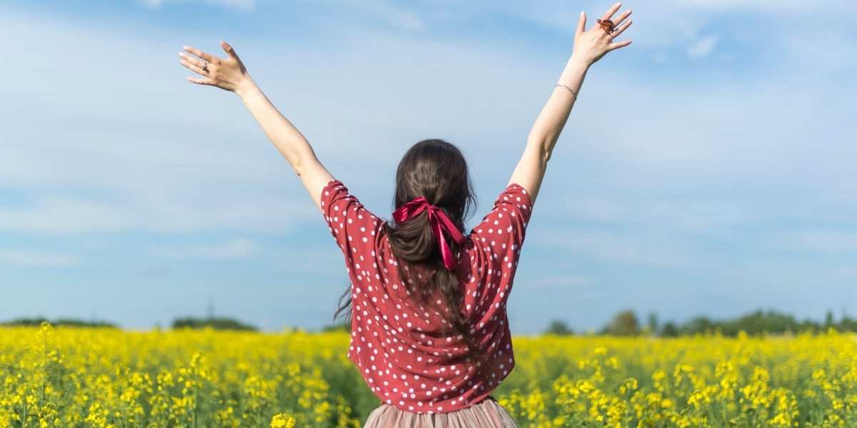A woman in a polka dot shirt raising her arms in a bright yellow flower field, expressing freedom and joy.