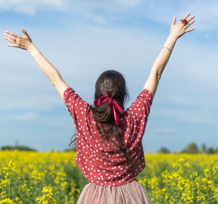 A woman in a polka dot shirt raising her arms in a bright yellow flower field, expressing freedom and joy.
