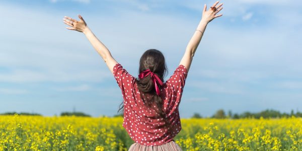 A woman in a polka dot shirt raising her arms in a bright yellow flower field, expressing freedom and joy.